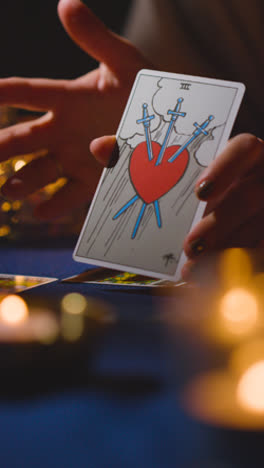 Vertical-Video-Close-Up-Of-Woman-Giving-Tarot-Card-Reading-On-Candlelit-Table-Holding-Card-Showing-Heart-Pierced-By-Swords